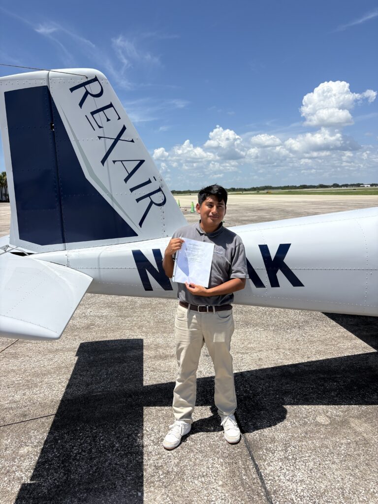 Young man in gray polo stands beside a small white airplane with blue tail, holding papers outside on a sunny tarmac.",