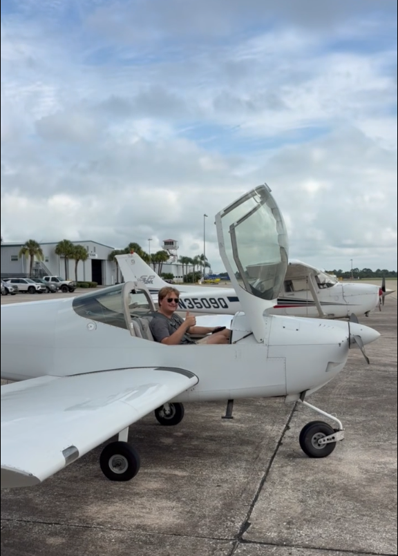 Person sits in a small white single-seat airplane with the cockpit canopy open on a runway, other planes and palm trees in the background.